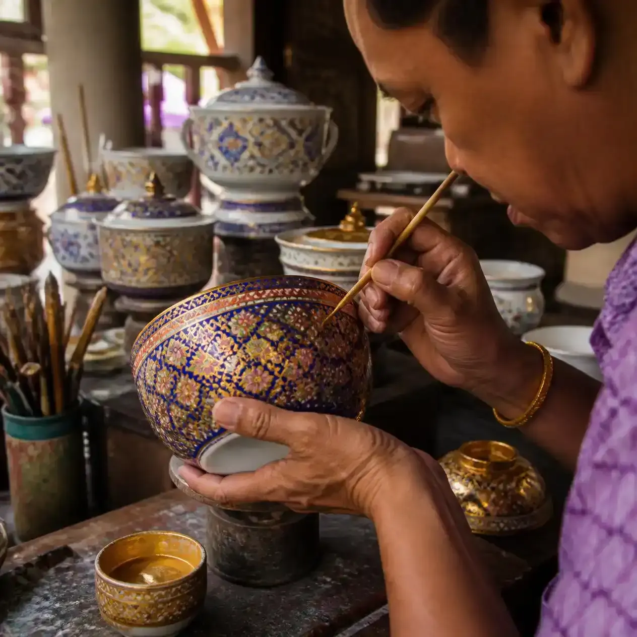 Thai artisan hand-painting fine gold detail onto a five-colour Benjarong porcelain bowl in a Bangkok workshop