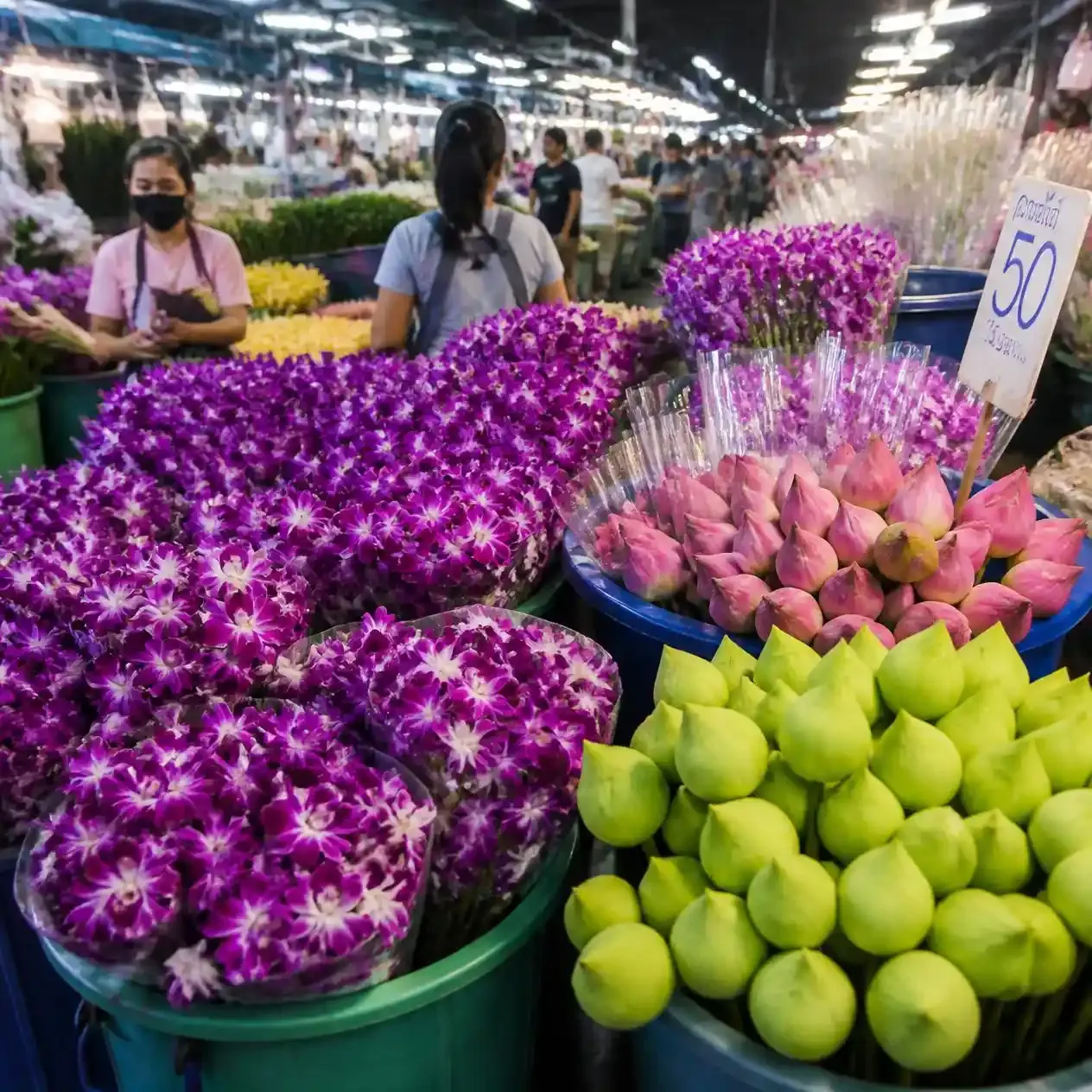 Buckets of purple orchids and lotus buds under strip lighting inside Pak Khlong Talat flower market in Bangkok
