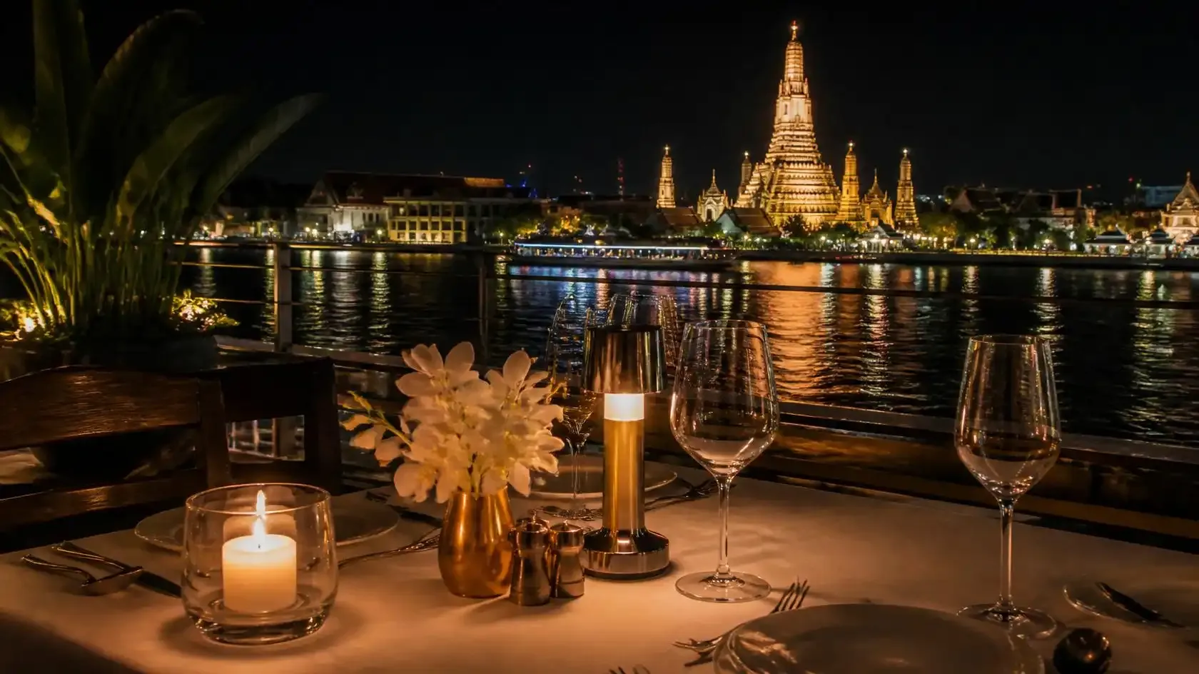 Candlelit table on a narrow riverside restaurant terrace in Bangkok with the Chao Phraya River and a floodlit temple behind