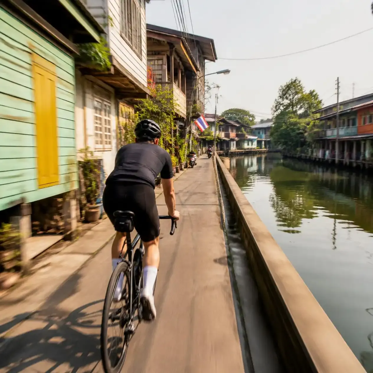Cyclist riding through a narrow Thonburi lane past wooden shophouses beside a canal in Bangkok