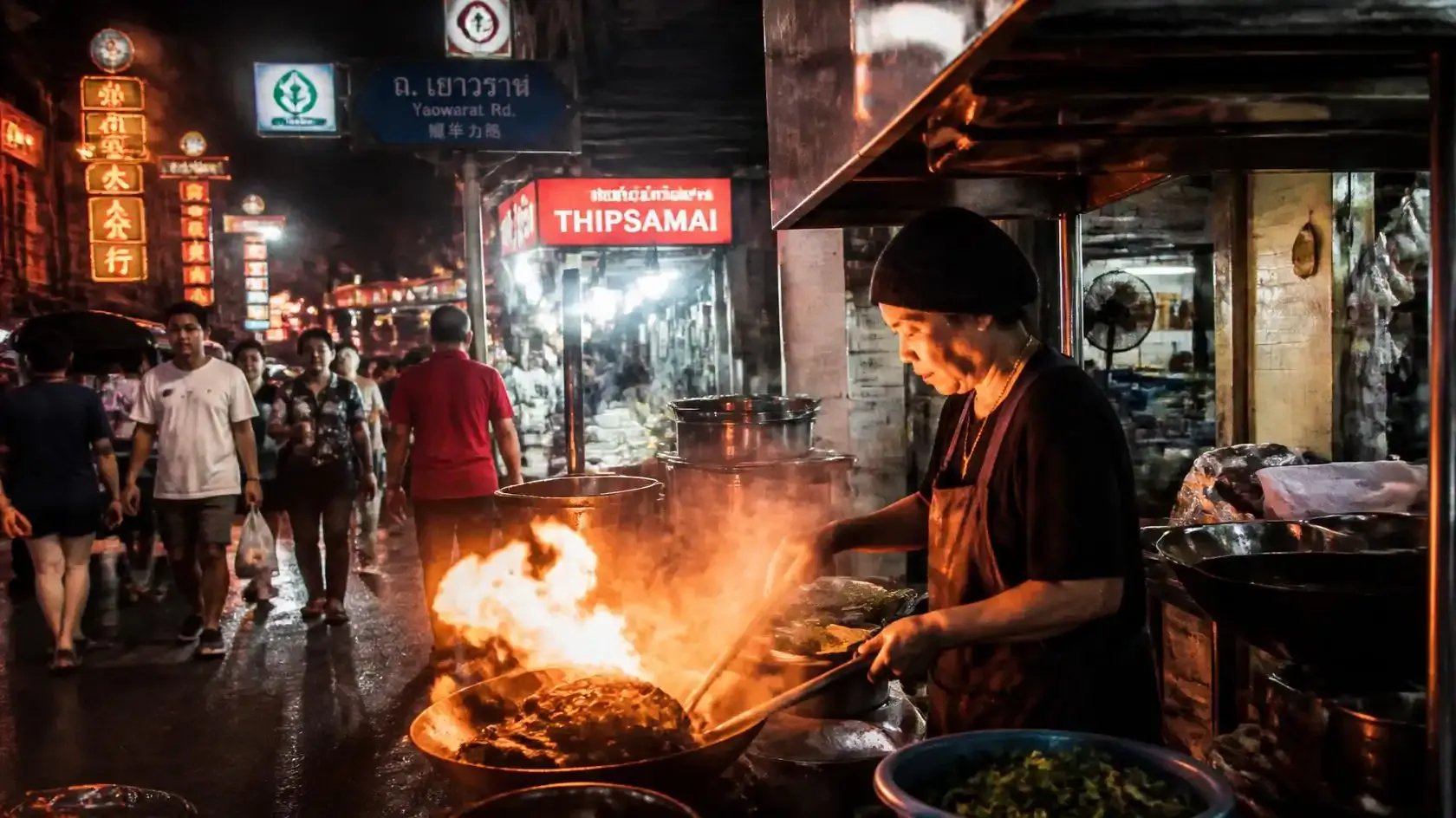 Cook tossing noodles over an open charcoal flame at a night-time street food stall on Yaowarat Road in Bangkok