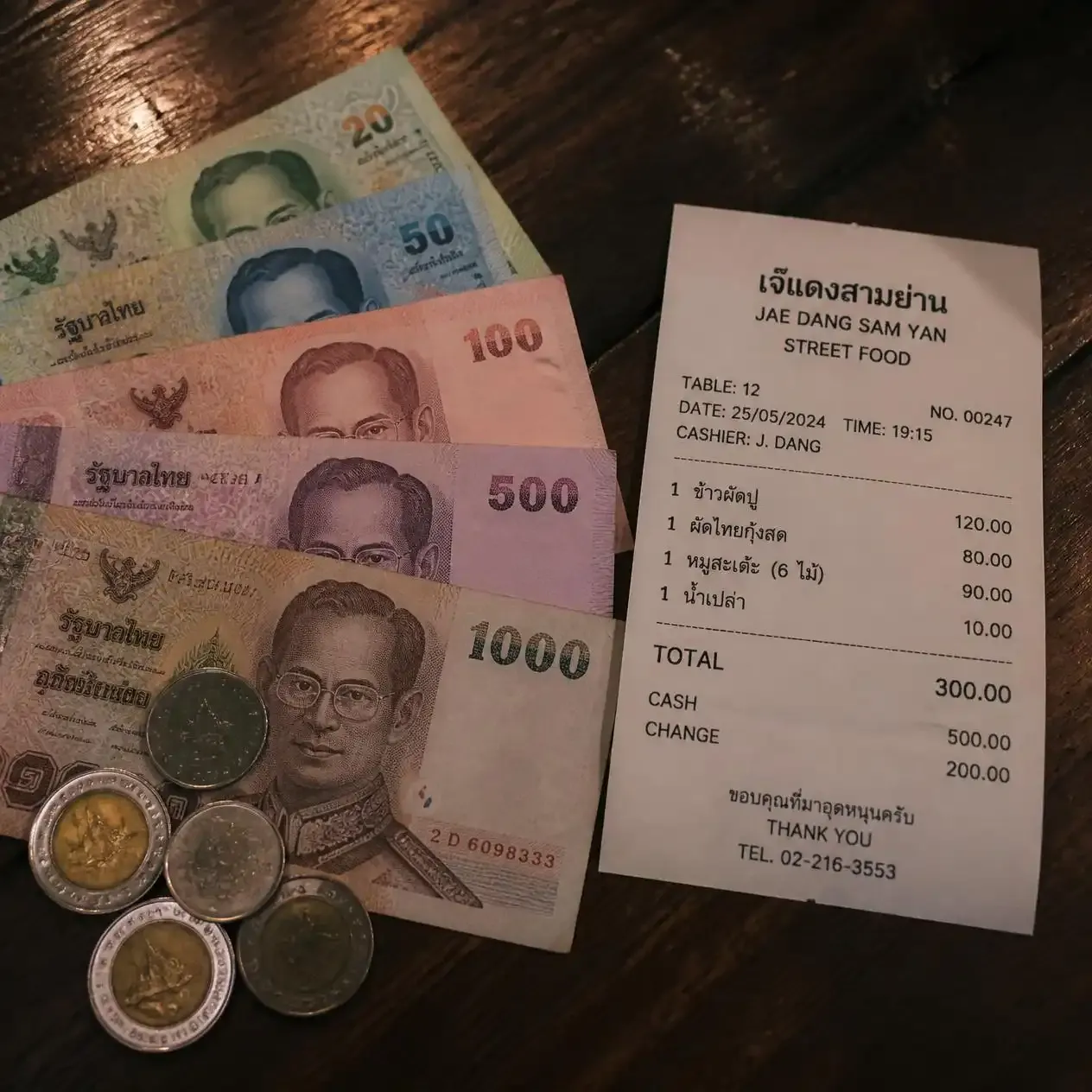 Thai baht banknotes and coins spread on a wooden table next to a street food receipt in Bangkok