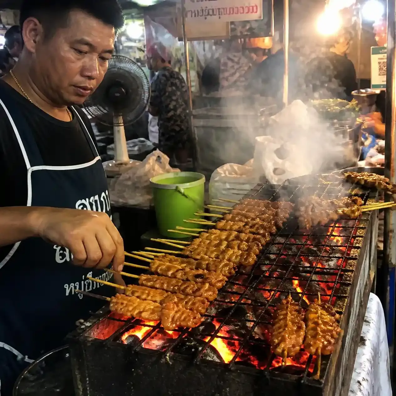 Vendor turning marinated pork skewers over a charcoal grill, a staple of street food in Bangkok