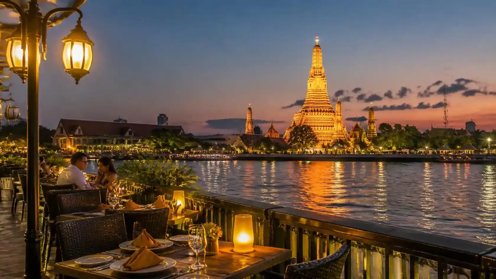 Wat Arun temple lit at dusk viewed across the Chao Phraya River from a candlelit riverside restaurant terrace in Bangkok