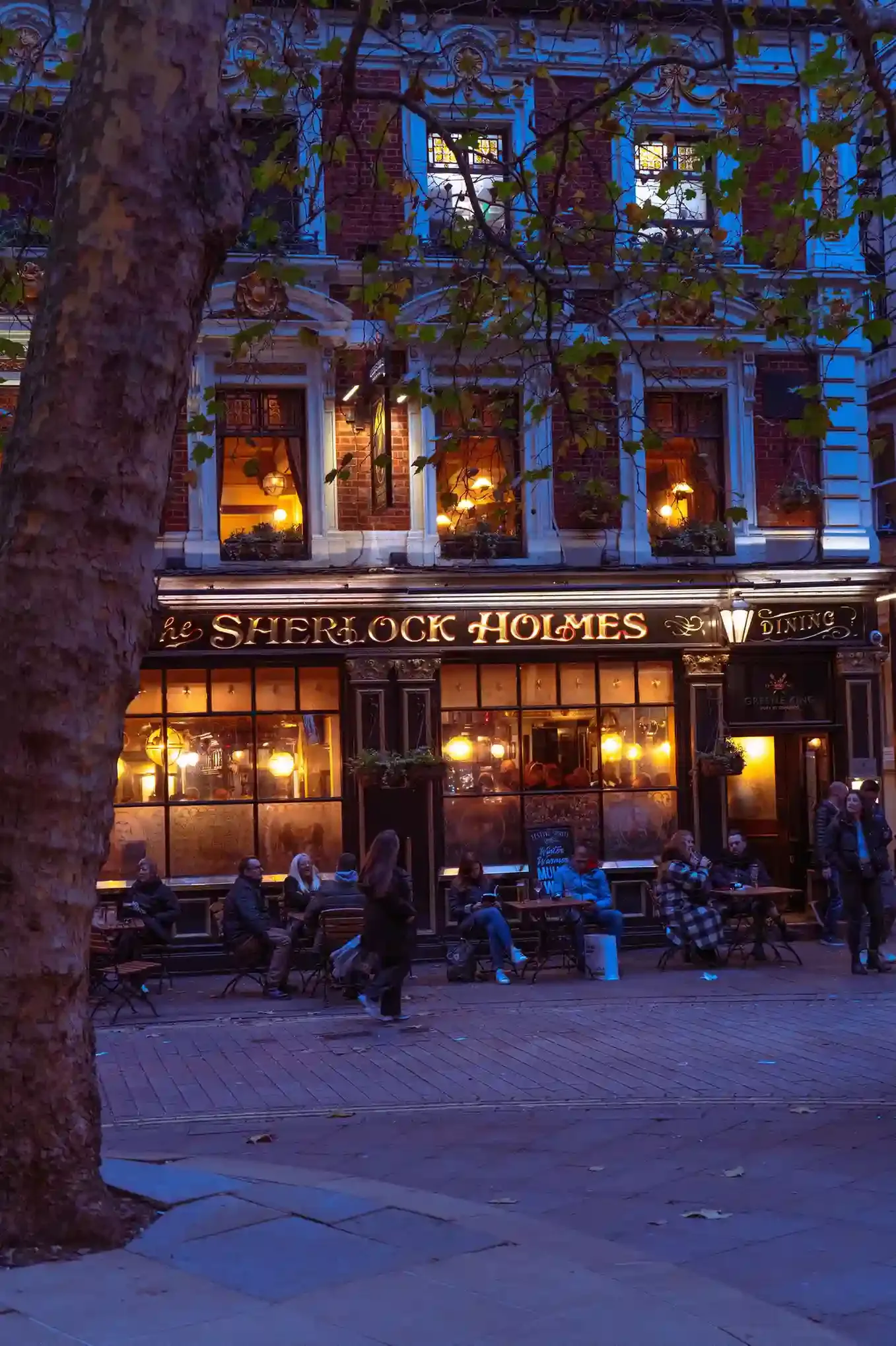 Diners at pavement tables outside busy restaurants on a lit Soho street in the London evening